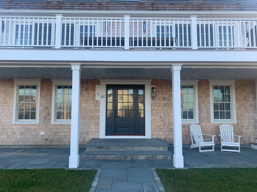 Front view of a coastal home with a porch, stone steps, and white lawn chairs.