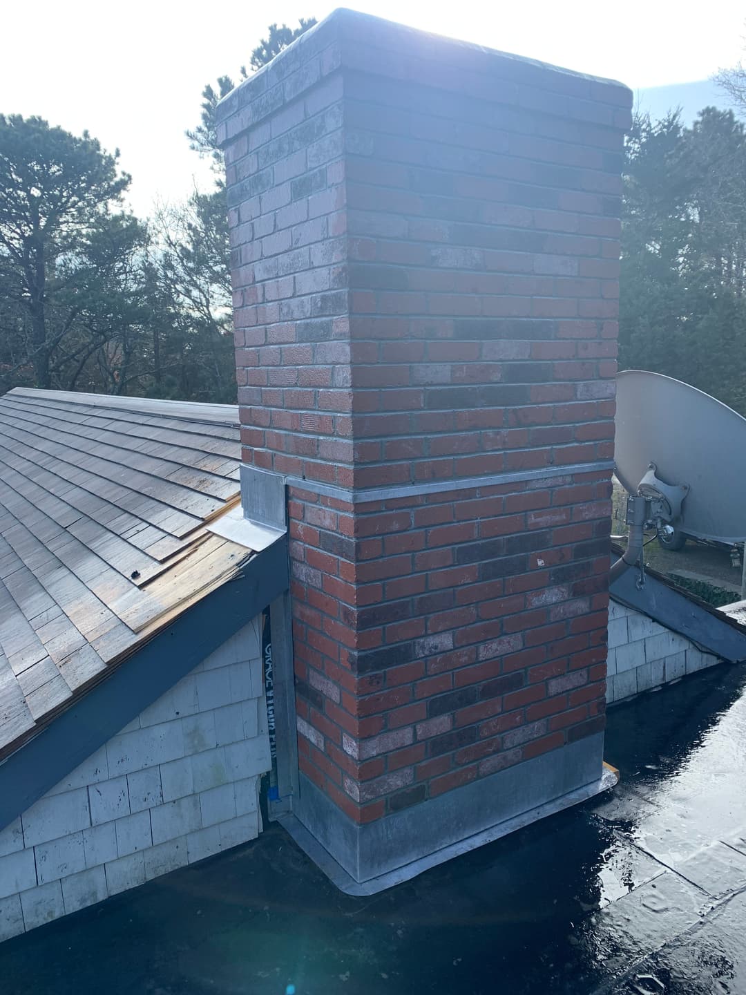 Brick chimney on a sloped roof with a satellite dish and trees in the background.