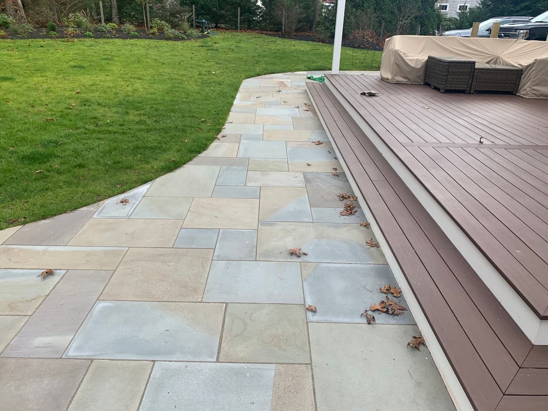 Paved walkway leading to a wooden deck in a landscaped yard with green grass.