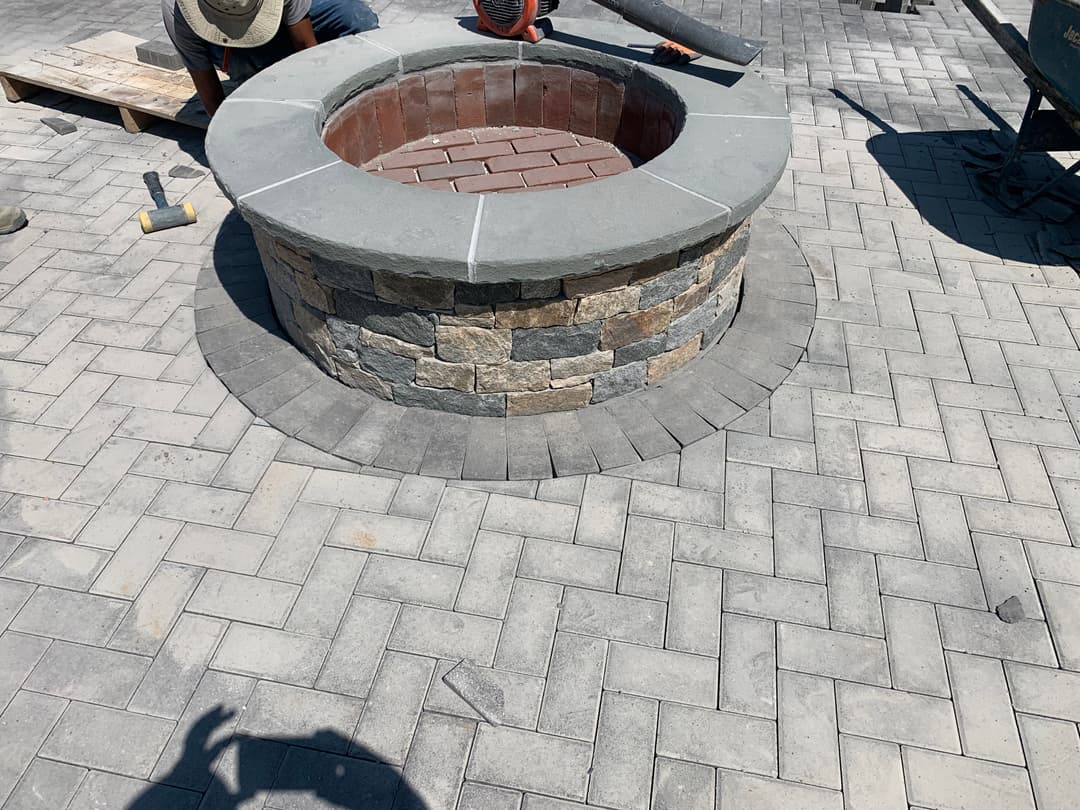 Stone fire pit construction on a brick patio with workers in the background.