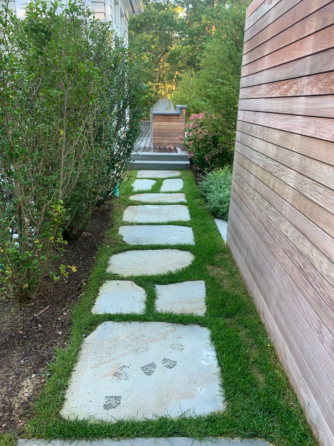 Stone pathway with grass and shrubs leading to a wooden deck in a landscaped garden.