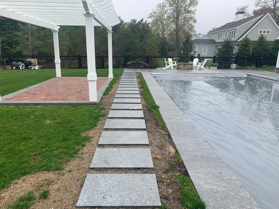 Stone walkway leads to a pool area with a covered patio and green lawn.
