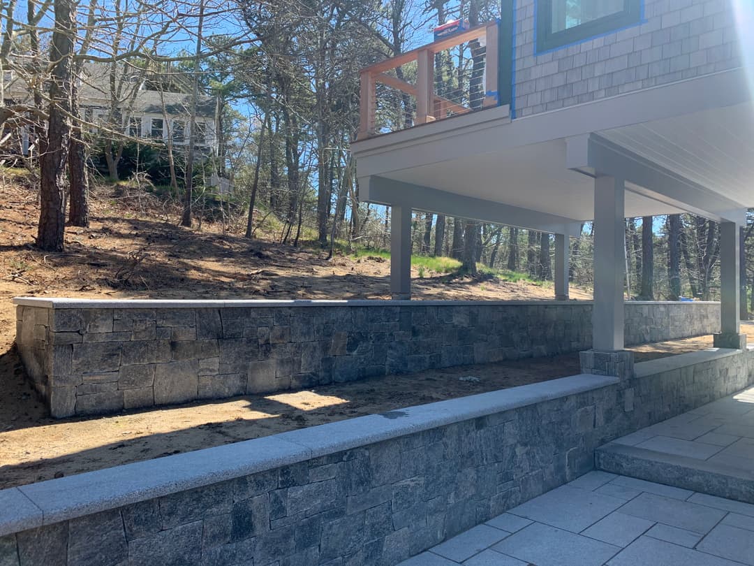 Stone retaining wall with patio area beneath a modern house in a wooded setting.