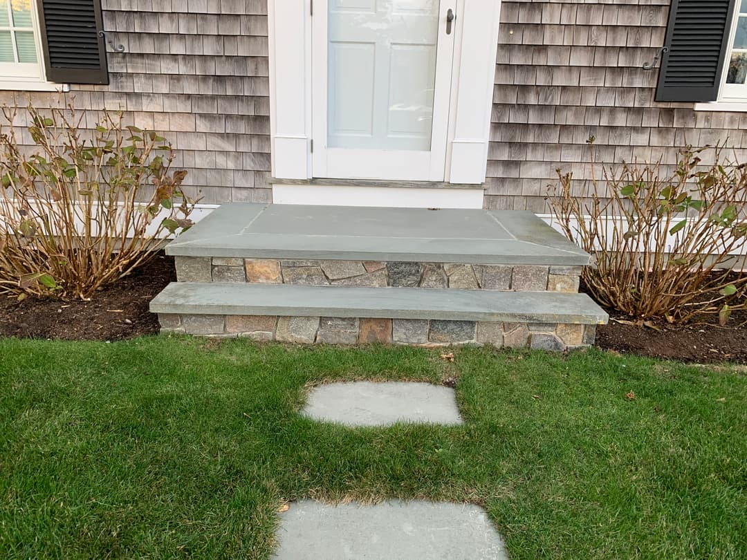 Stone front steps leading to a white door, surrounded by shrubs and a green lawn.