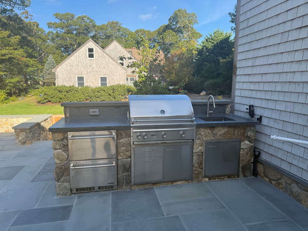 Outdoor kitchen with stainless steel grill and stone countertop, set in a landscaped yard.