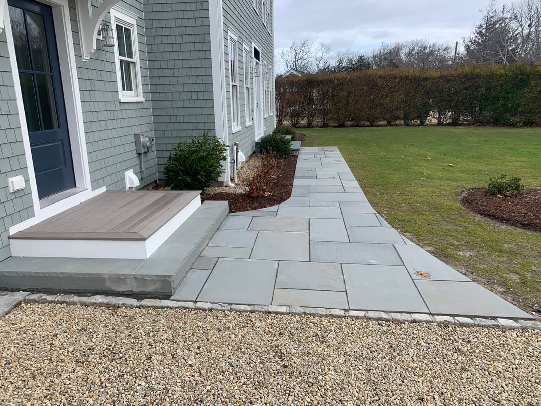 Stone pathway leading to a blue house with a porch, surrounded by a landscaped yard.