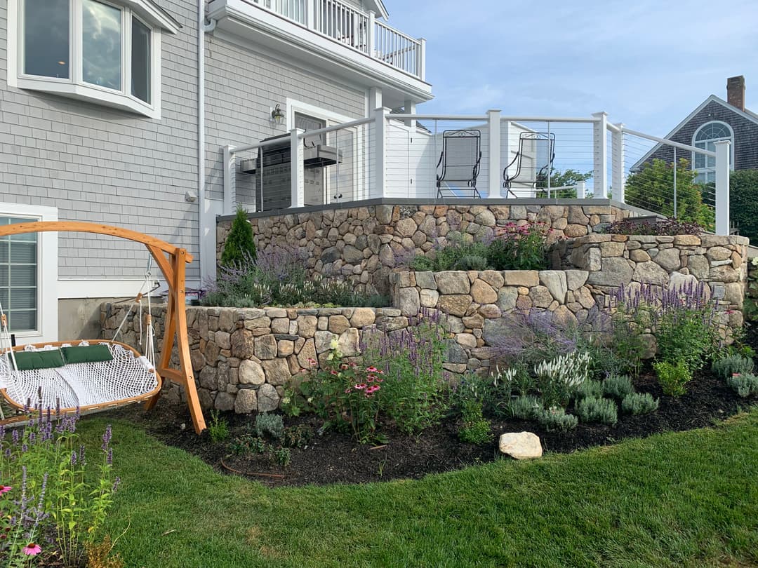 Landscape design featuring stone terrace, flowering plants, and a hanging swing by a house.
