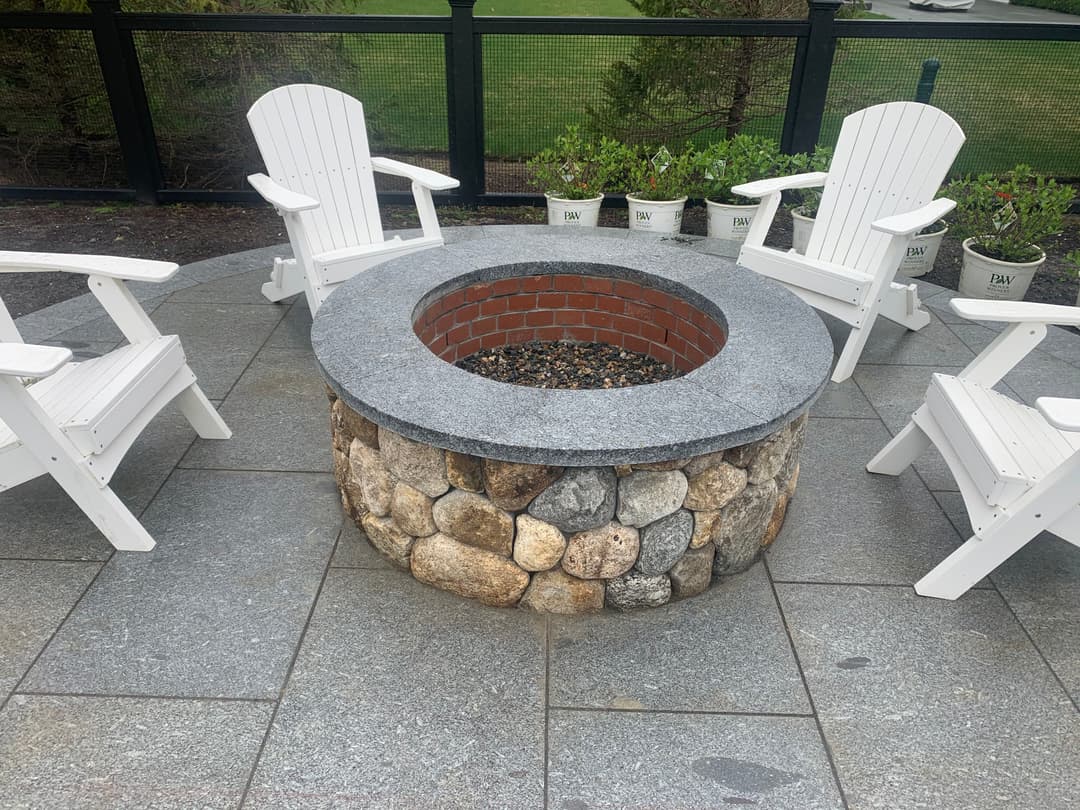 Stone fire pit surrounded by white patio chairs, set in a landscaped outdoor area.