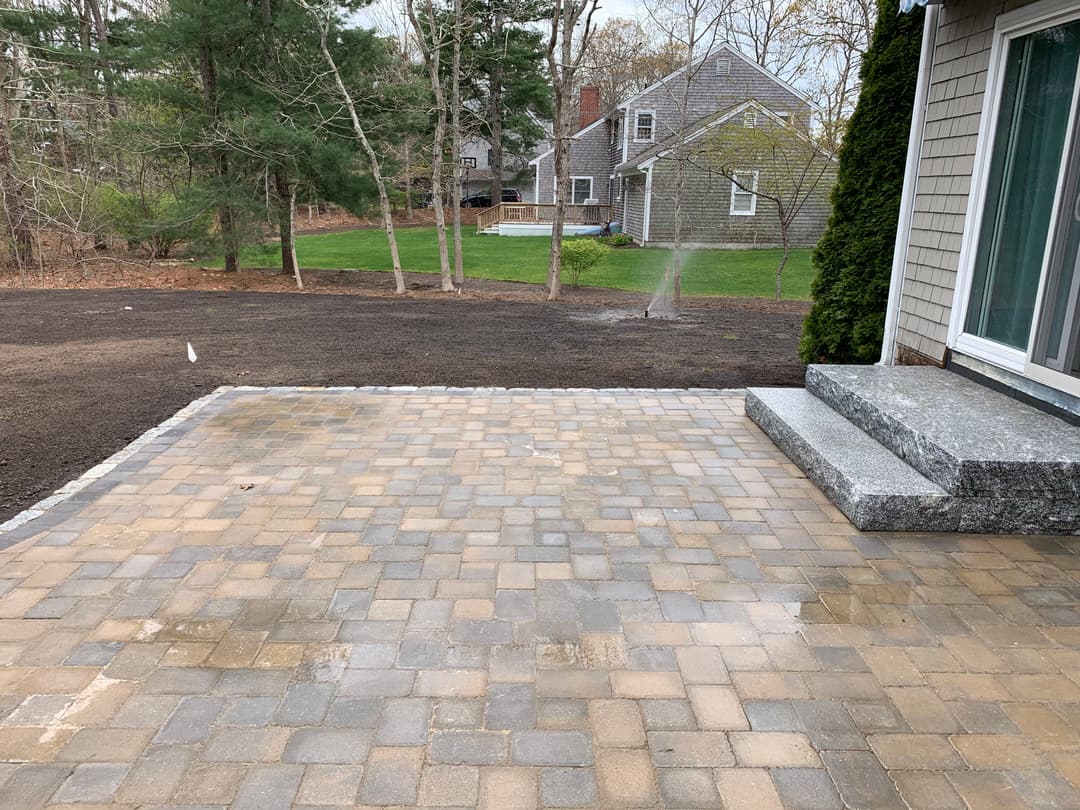 Paver patio with stone steps overlooking a grassy backyard and wooded area.