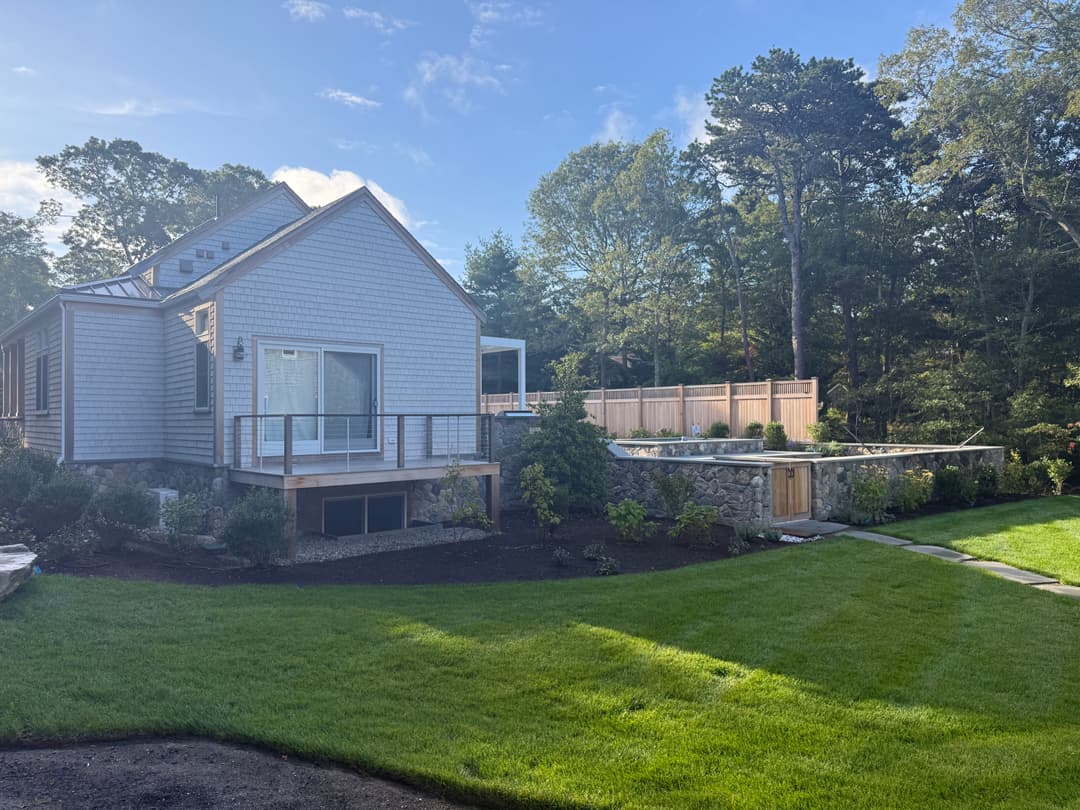 Modern gray house with landscaped yard and stone wall, surrounded by trees under a bright sky.