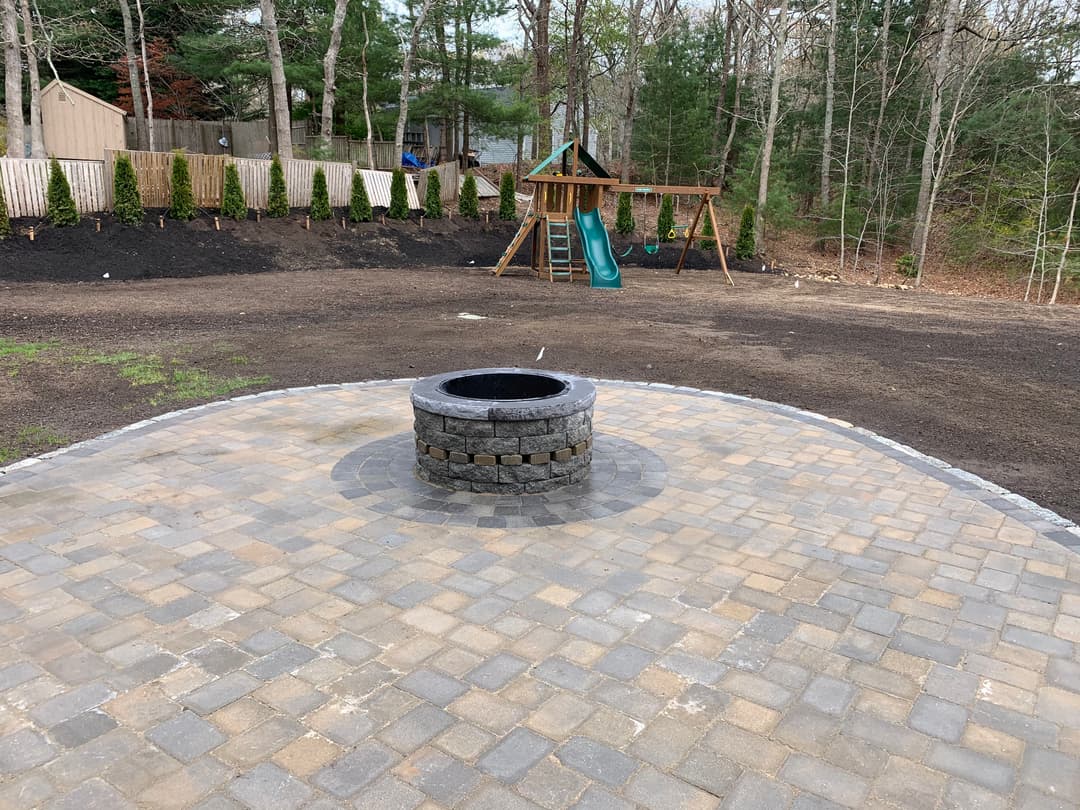 Backyard patio with a stone fire pit and playground set in a wooded setting.