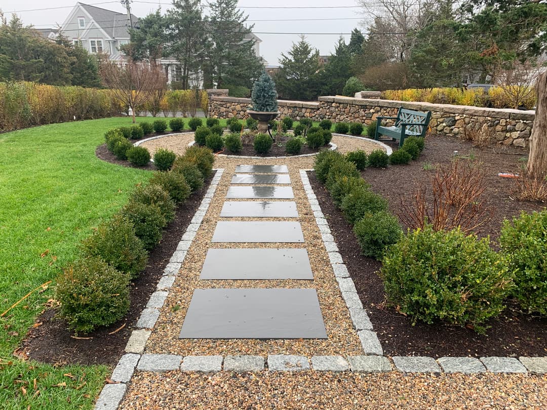 Landscaped garden pathway with stepping stones, shrubs, and stone wall backing.