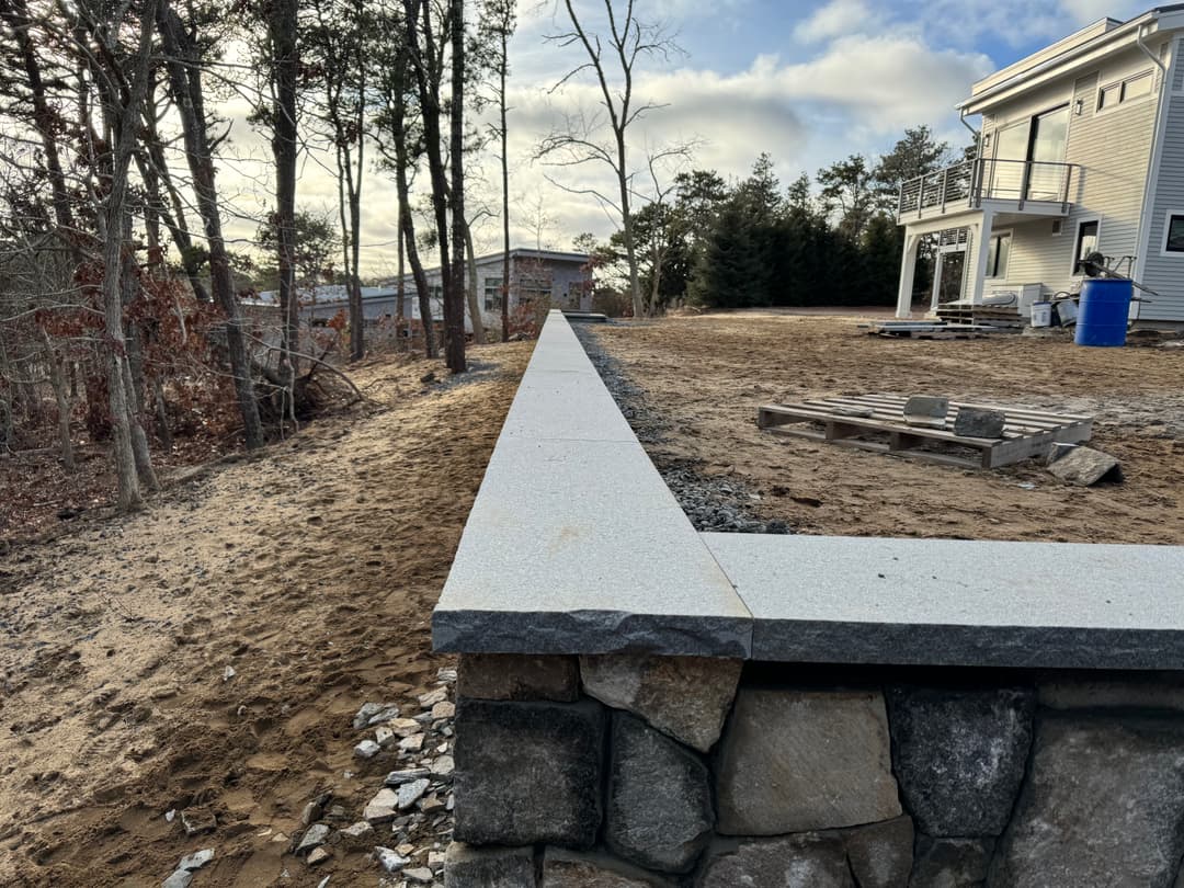 Stone retaining wall with a concrete cap near a construction site in a wooded area.