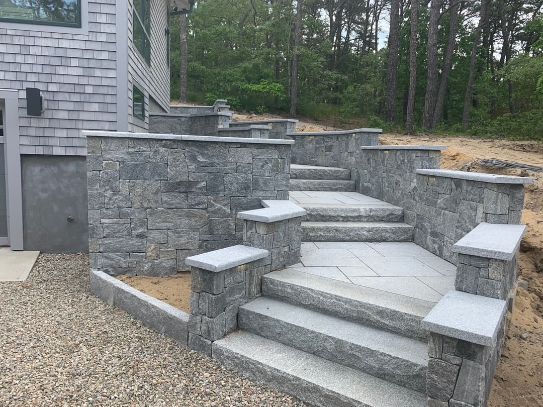 Stone outdoor stairs with retaining walls leading to a house in a wooded area.
