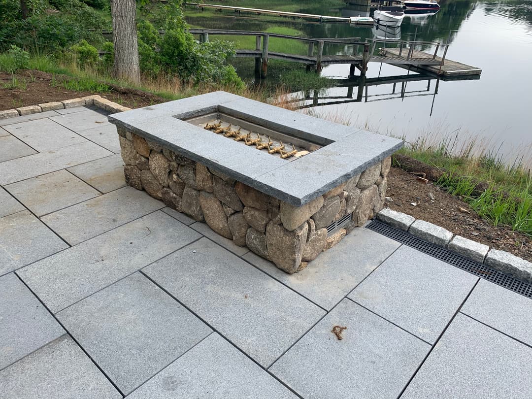 Outdoor stone fire pit with granite top by a calm lake, surrounded by patio pavers.