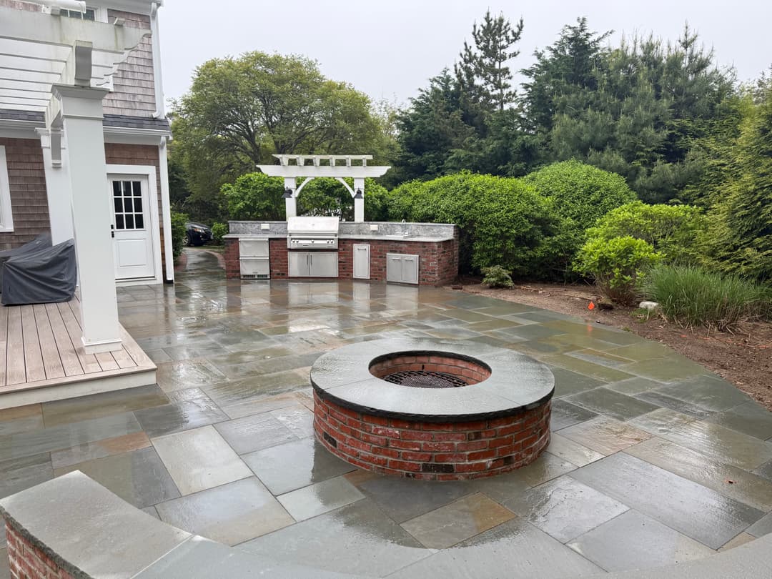 Backyard patio with stone landscaping, built-in grill, and circular fire pit surrounded by greenery.
