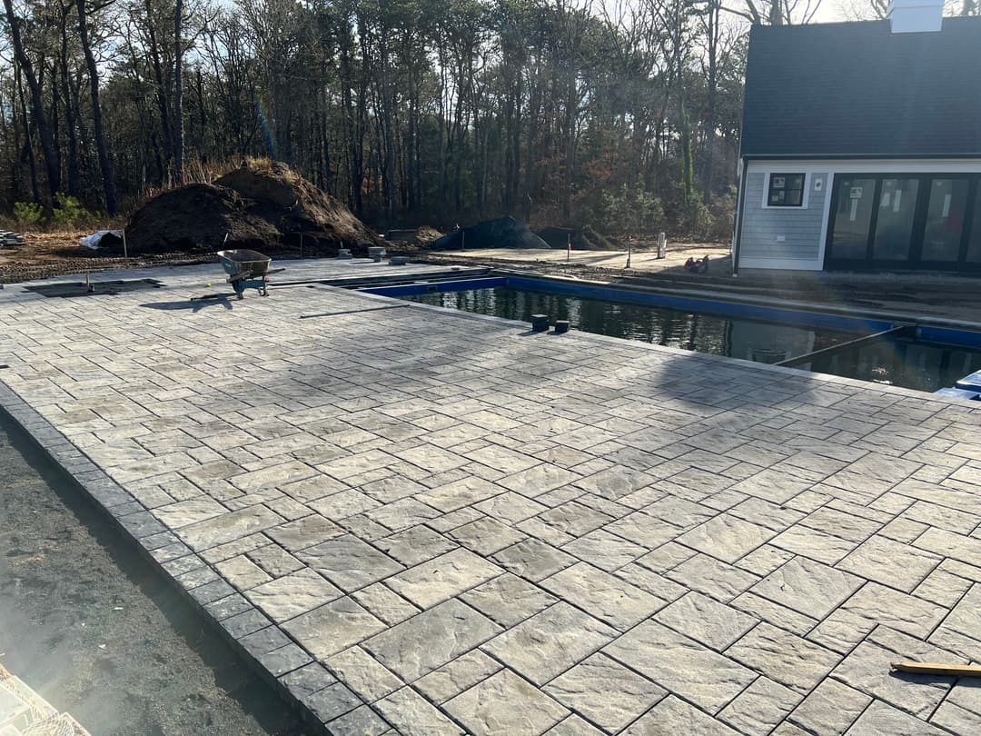 Stone patio installation near a pool in a wooded residential area.