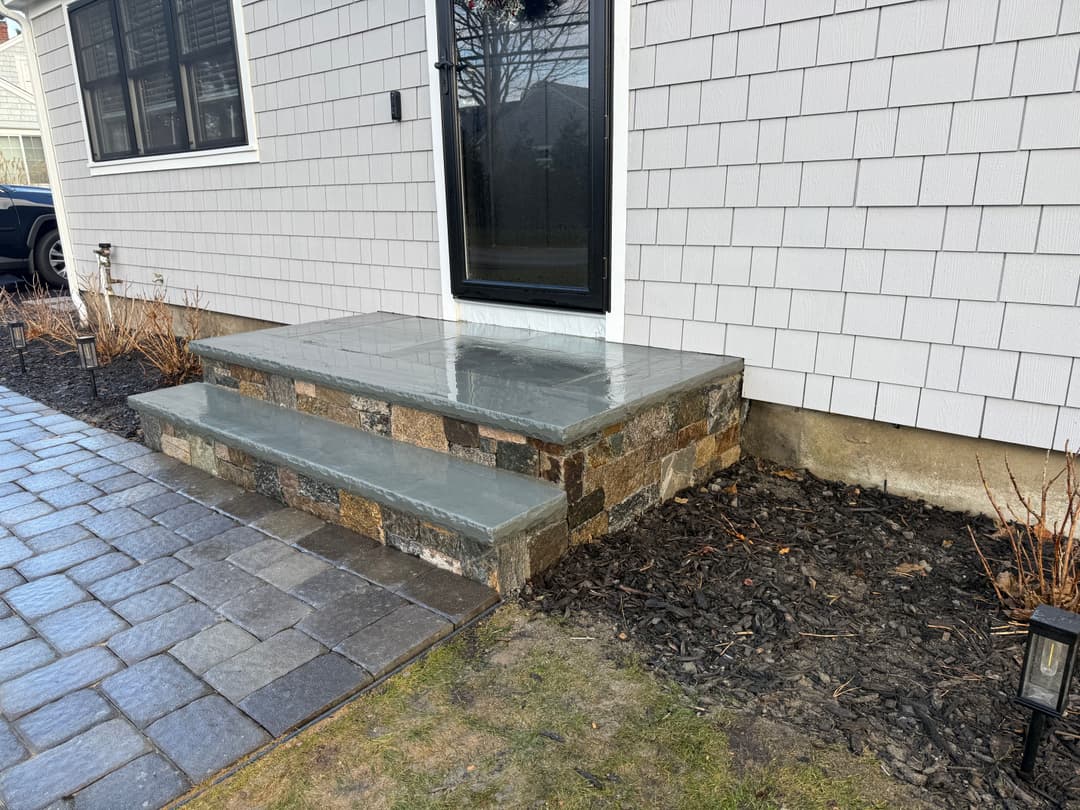 Front entrance with stone steps, gray patio, and landscaped border, featuring a black door.