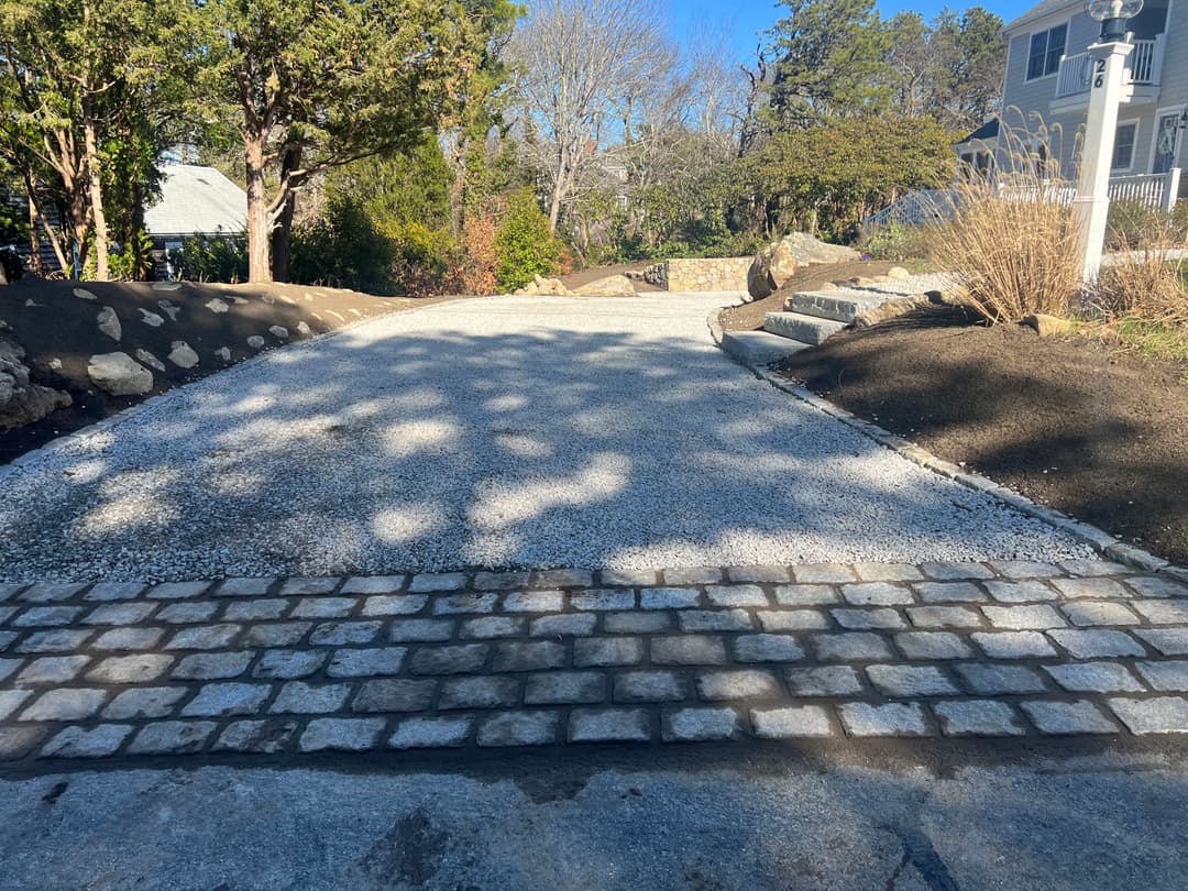 Gravel driveway with cobblestone borders, surrounded by landscaped greenery and trees.