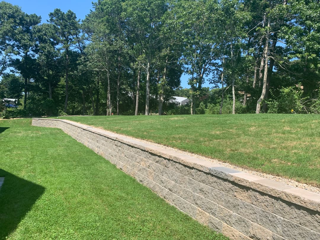 Stone retaining wall in a grassy landscape surrounded by trees under a clear blue sky.