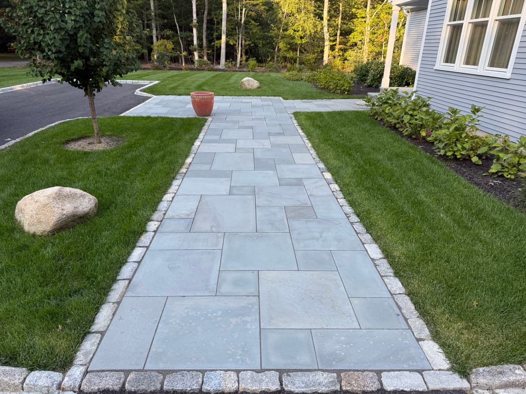 Stone walkway lined with grass and landscaping leading to a house in a serene setting.