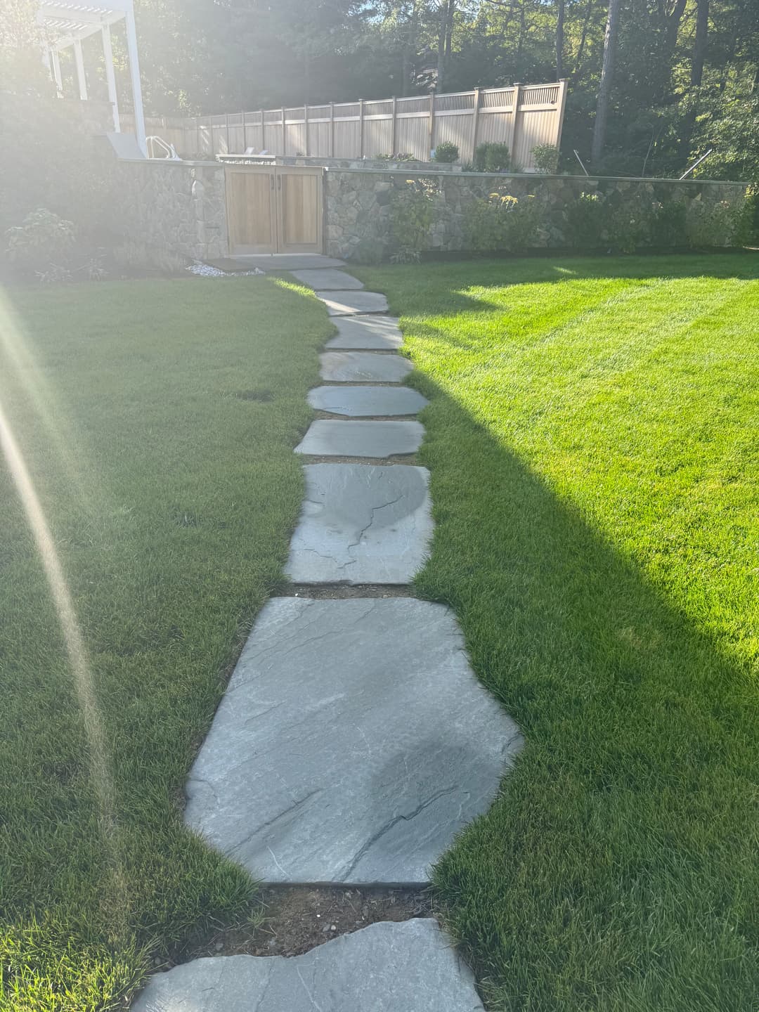 Stone pathway leading through lush green grass to a wooden gate surrounded by trees.