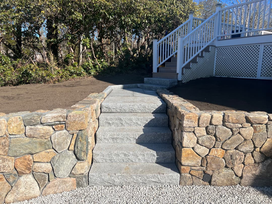 Granite steps lead up to a deck, surrounded by stone walls and landscaped greenery.