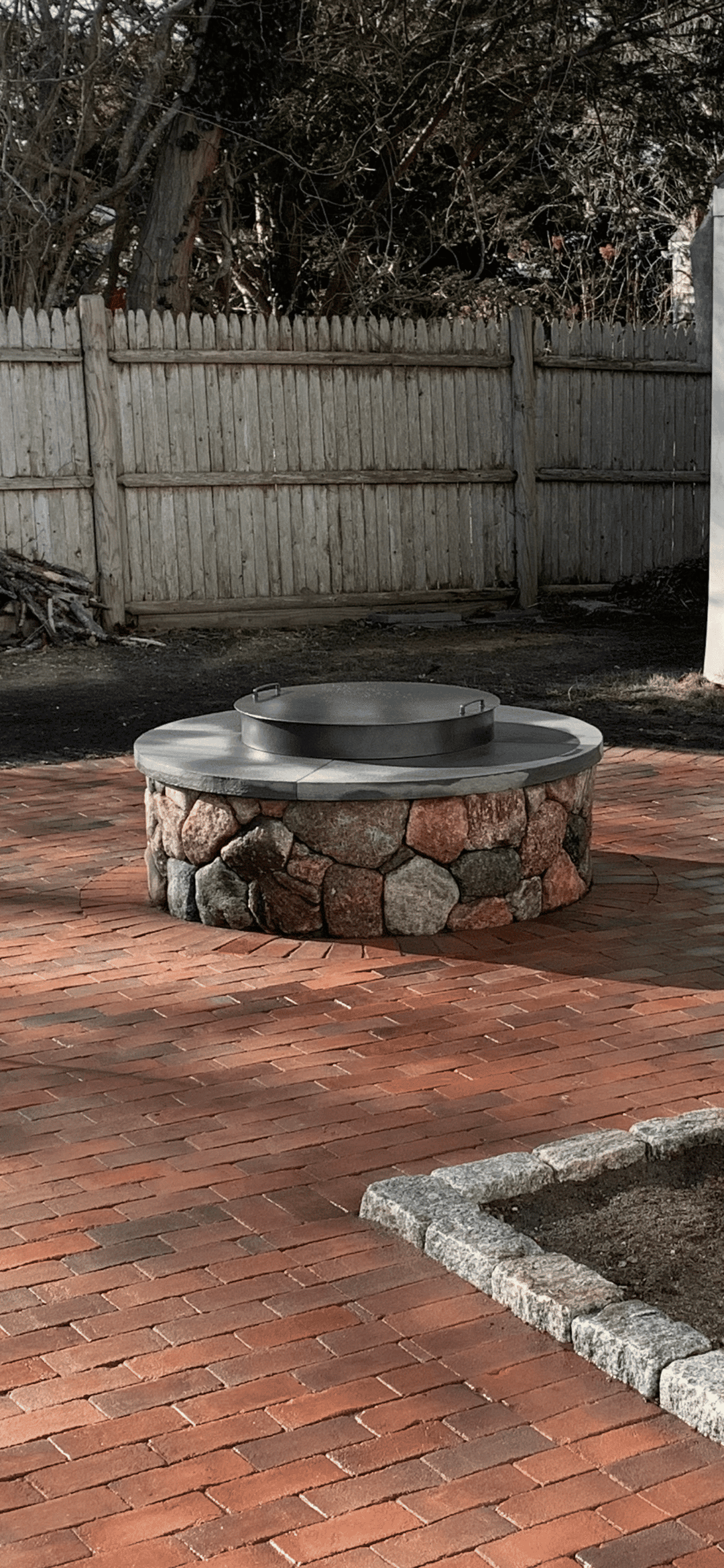 Round stone fire pit on a brick patio with a wooden fence in the background.