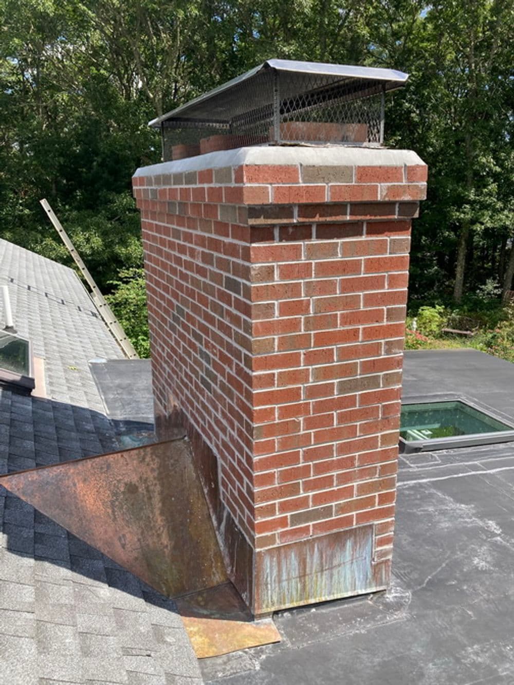 Chimney with brick construction and metal cap on a roof surrounded by trees.