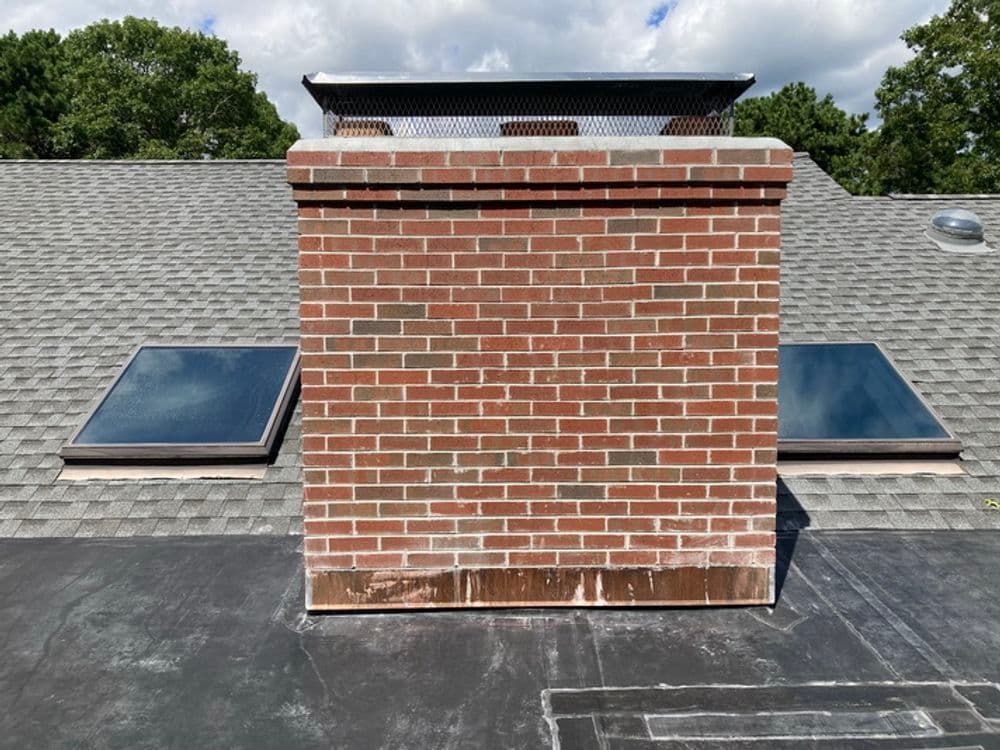 Brick chimney on a sloped roof with skylights and blue sky backdrop.