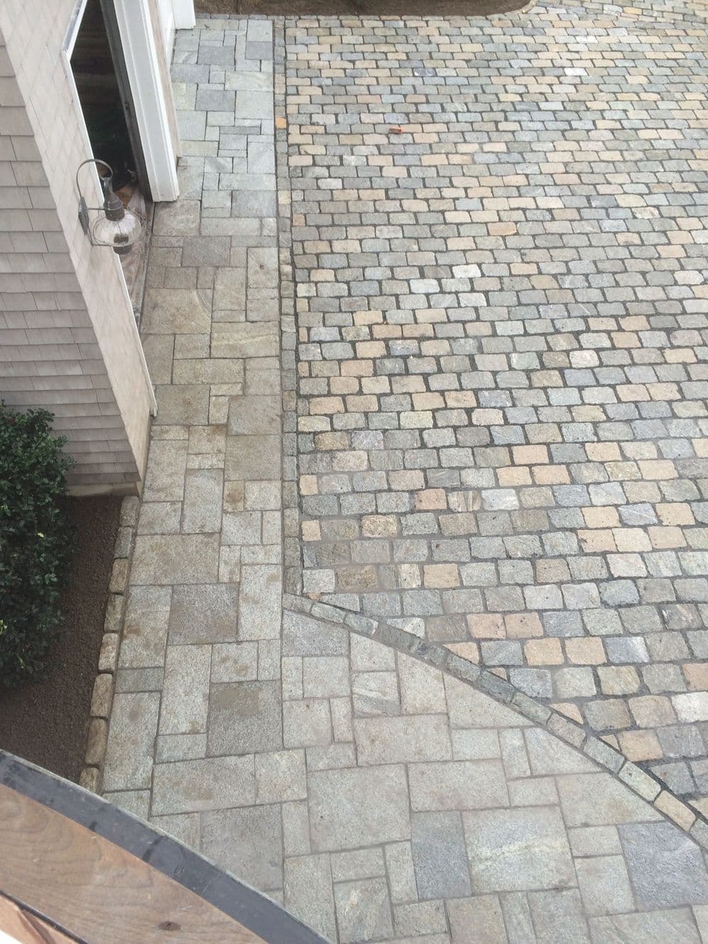 Paved stone driveway with intricate patterns, bordered by shrubs and a house exterior.
