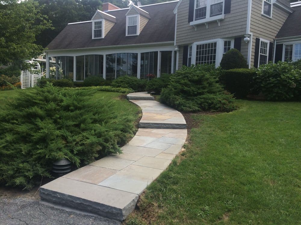 Curved stone pathway leading to a house surrounded by lush greenery and shrubs.