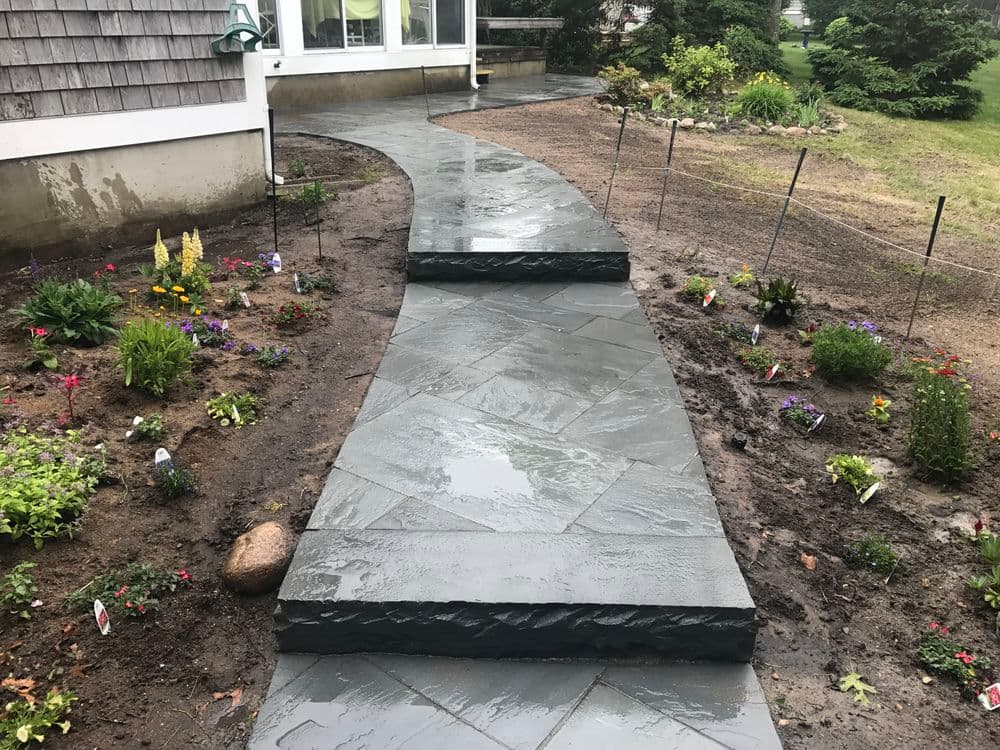 Wet stone pathway leads to a house, surrounded by colorful flower beds and greenery.