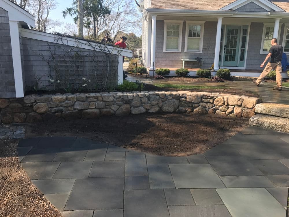 Stone wall and slate patio installation in front of a modern house with landscaping.