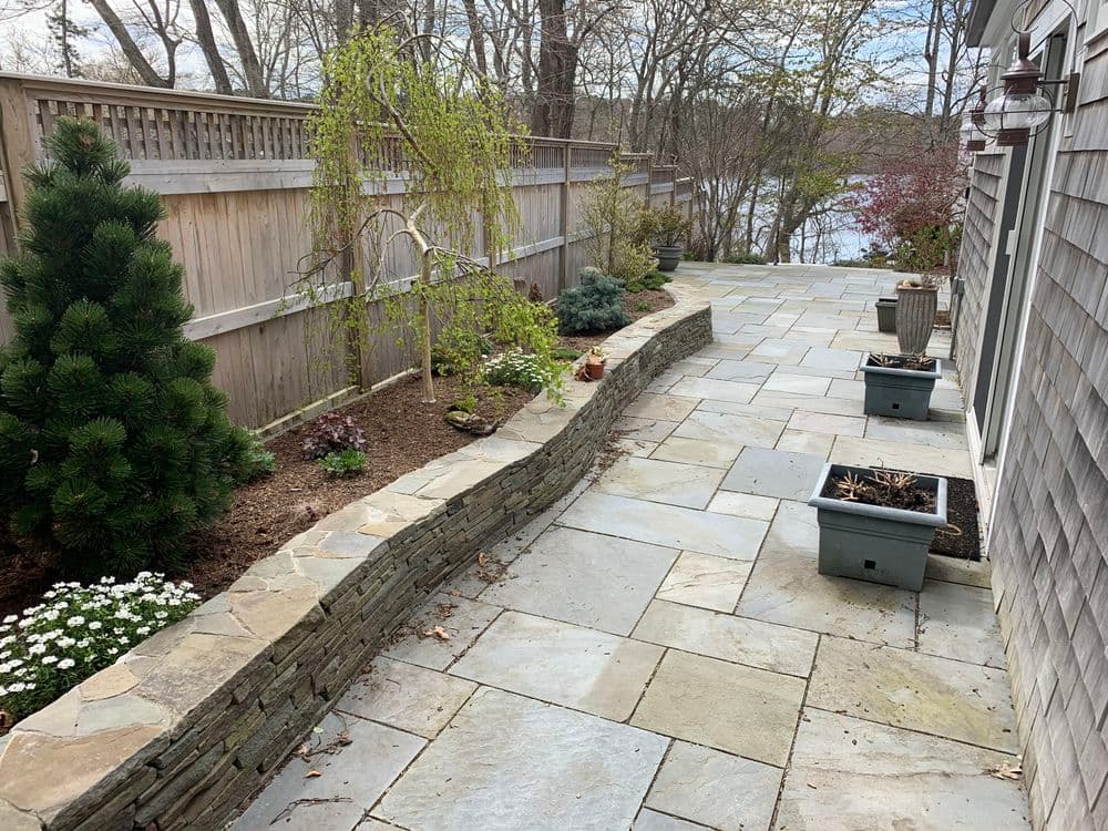 Stone patio with landscaped borders, planters, and trees near a serene lake view.
