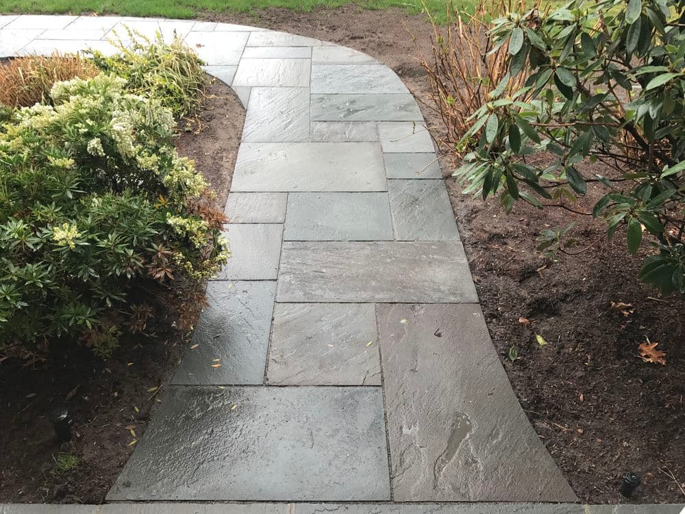 Paved walkway of dark stone curving through lush garden with plants and bushes.
