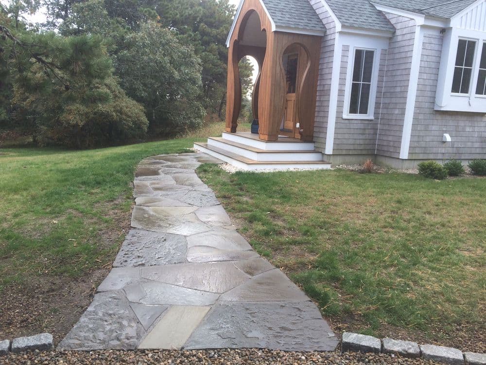 Stone pathway leading to a modern wooden entryway of a house surrounded by greenery.