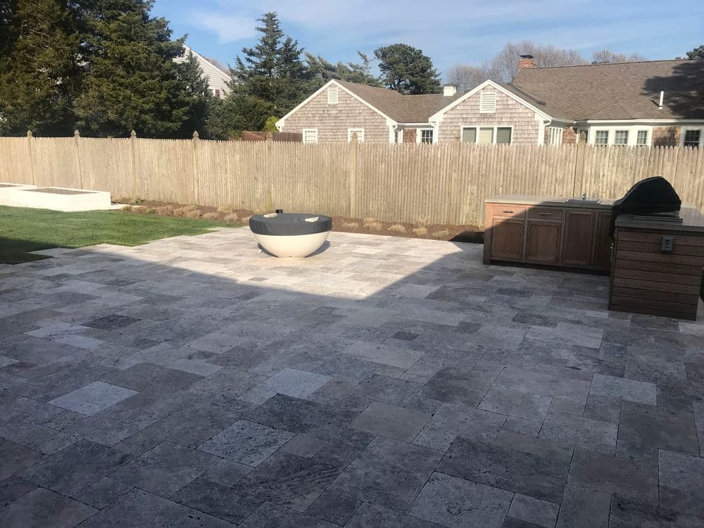 Modern outdoor patio with stone tiles, fire pit, and wooden cabinetry in a residential backyard.