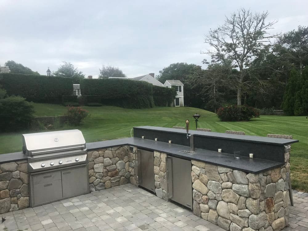 Outdoor kitchen with stone grill, stainless steel appliances, and manicured lawn backdrop.