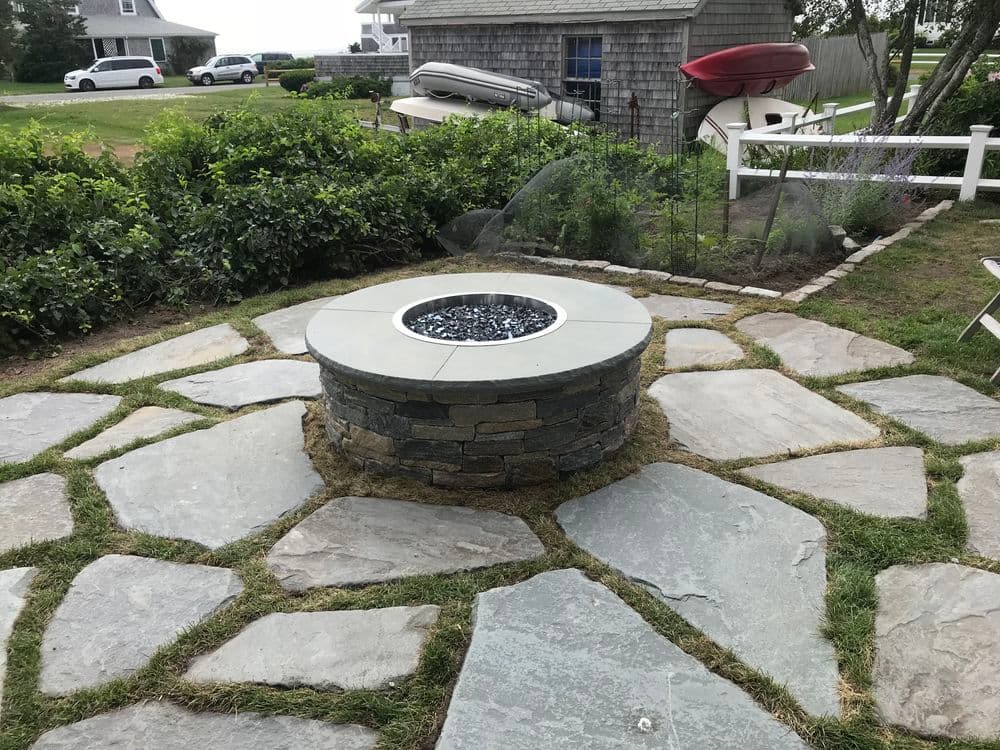 Outdoor stone fire pit surrounded by slate patio and lush greenery in coastal setting.