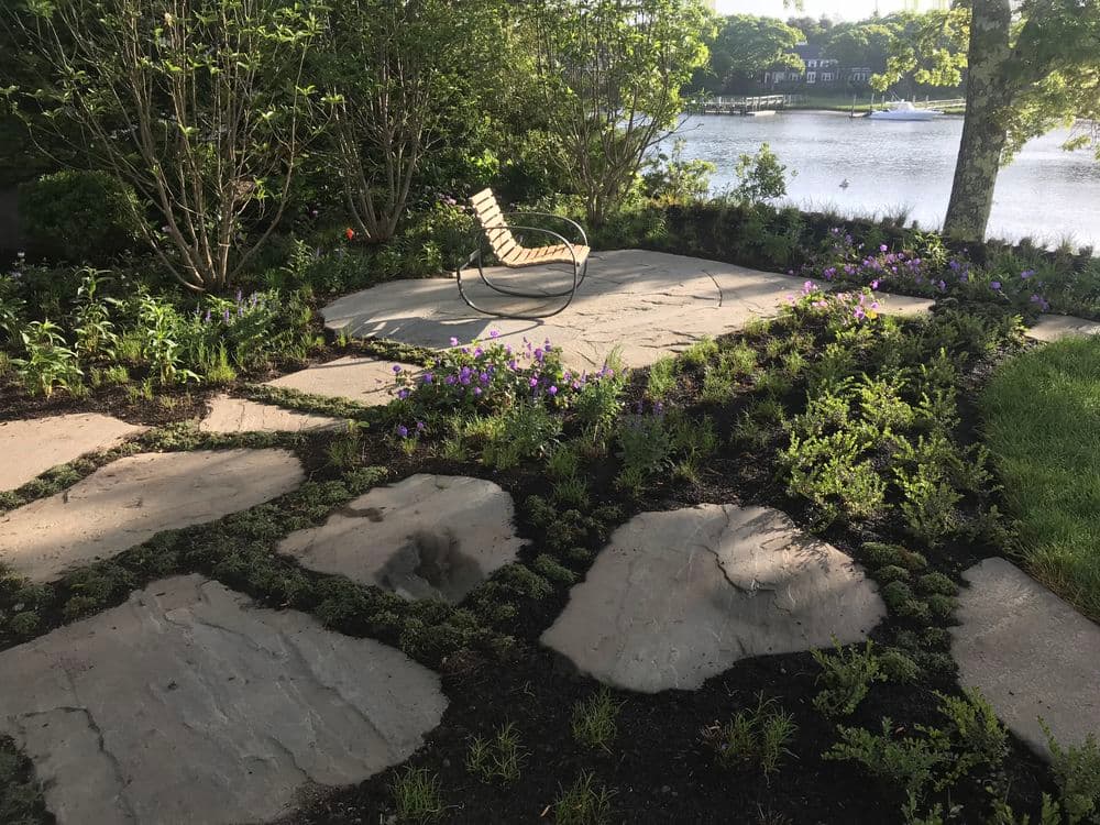 Lush garden patio with stone path, colorful flowers, and a relaxing chair by the water.