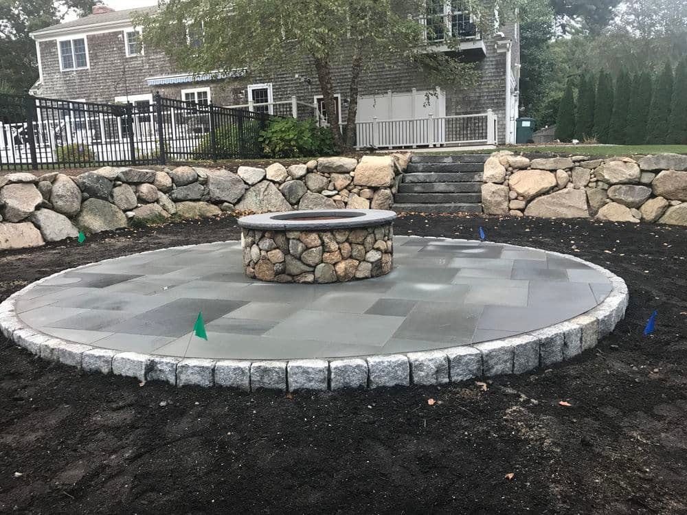 Patio with stone fire pit, surrounded by landscaping stones and grass area in backyard setting.