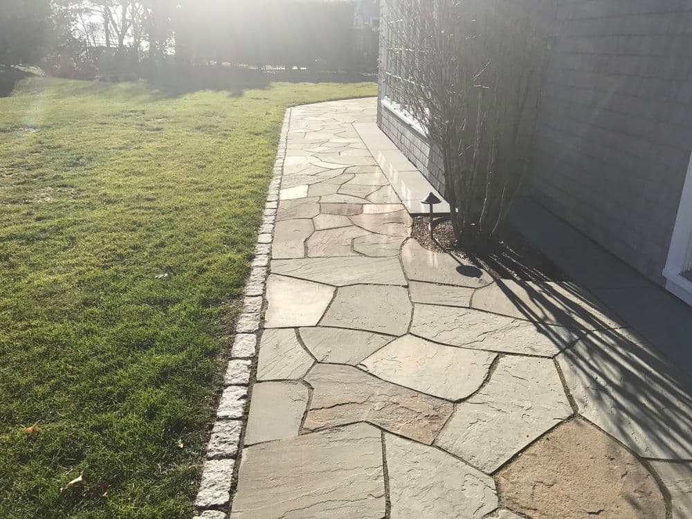 Natural stone pathway along a lawn, with sunlight casting shadows on a house wall.