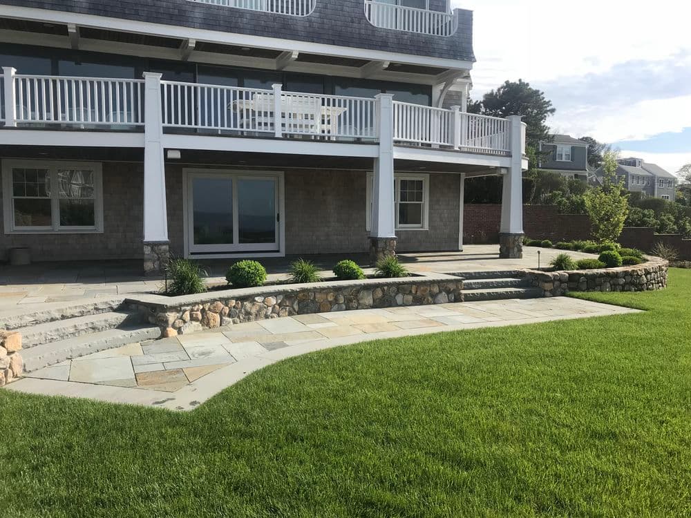 Coastal home with spacious patio, stone steps, and lush green lawn overlooking the water.