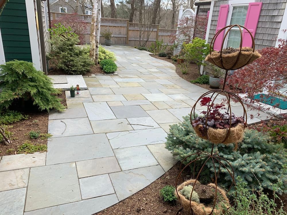 Stone patio path with garden greenery and a decorative plant stand in a serene outdoor setting.