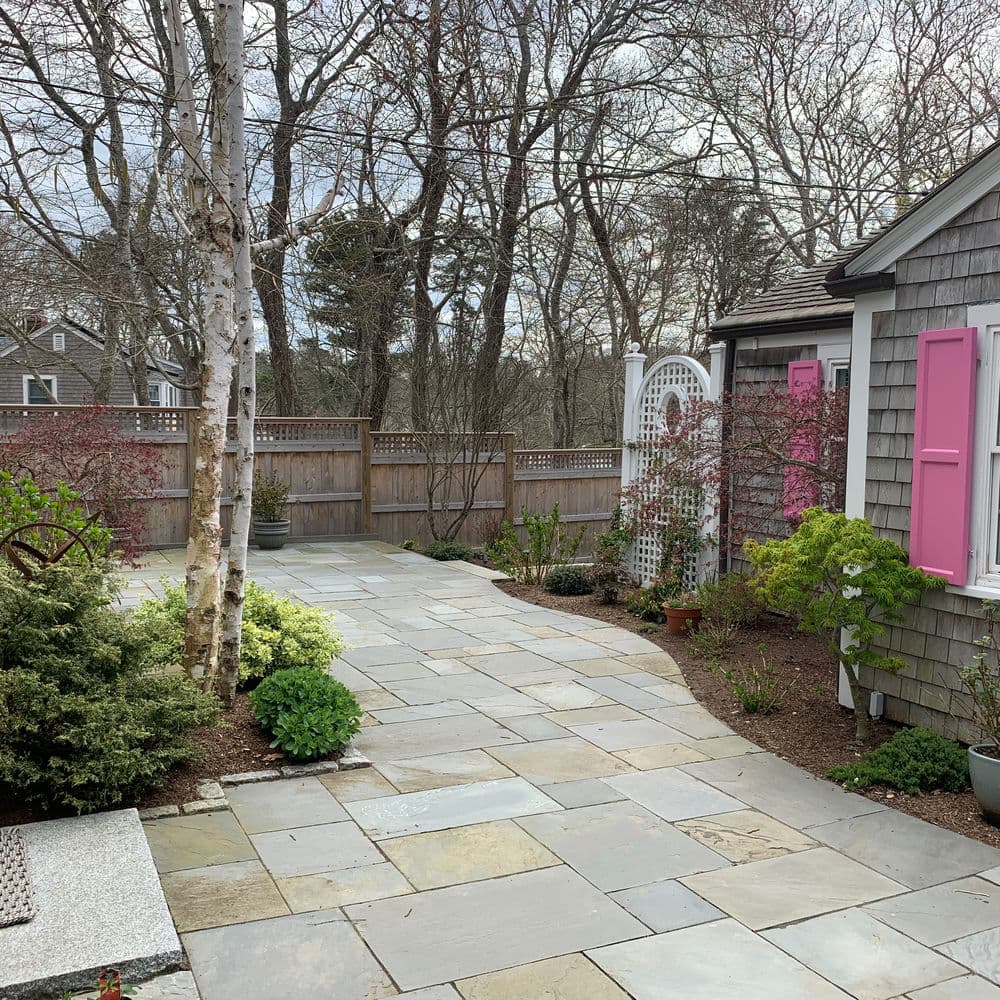 Paved garden pathway with colorful plants and a pink window, surrounded by trees and fencing.