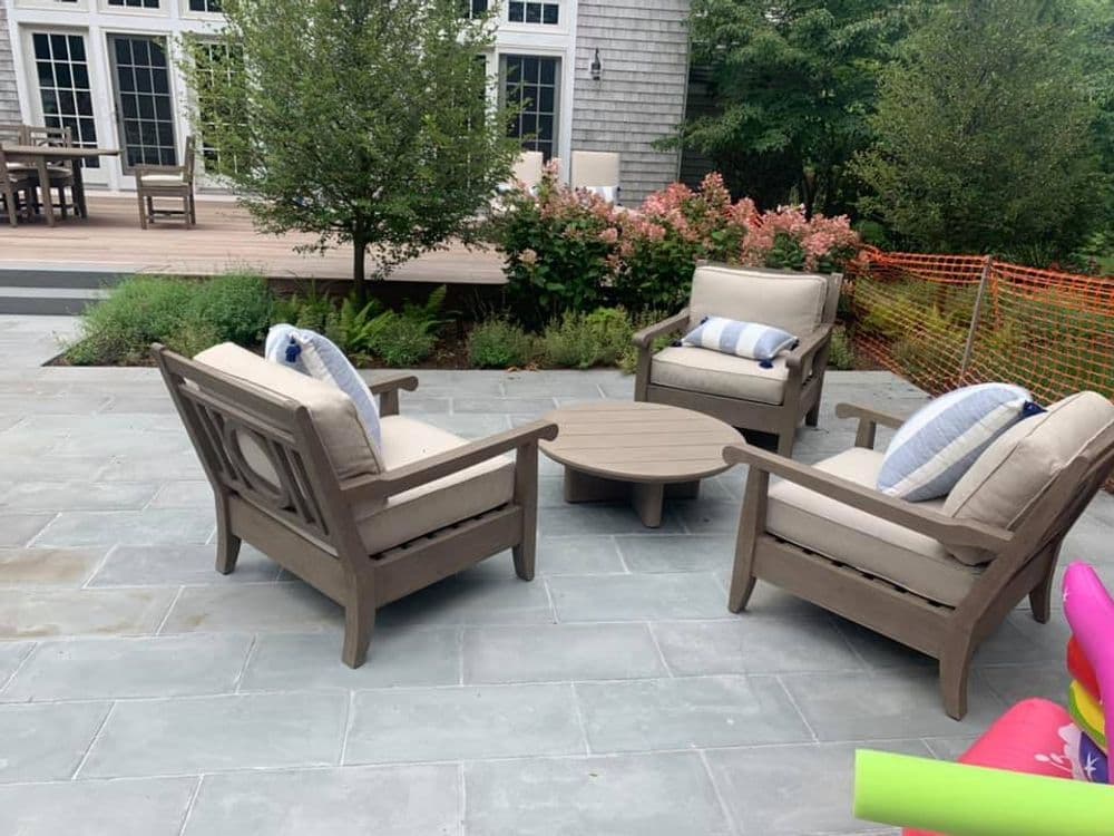 Outdoor seating area with four chairs and a round table on a stone patio surrounded by greenery.