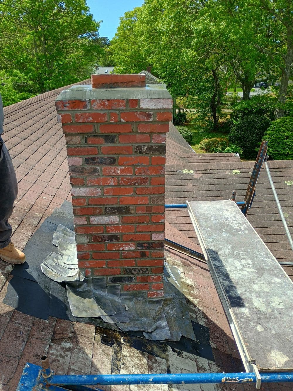 Chimney repair with brick structure on a rooftop surrounded by trees and scaffolding.