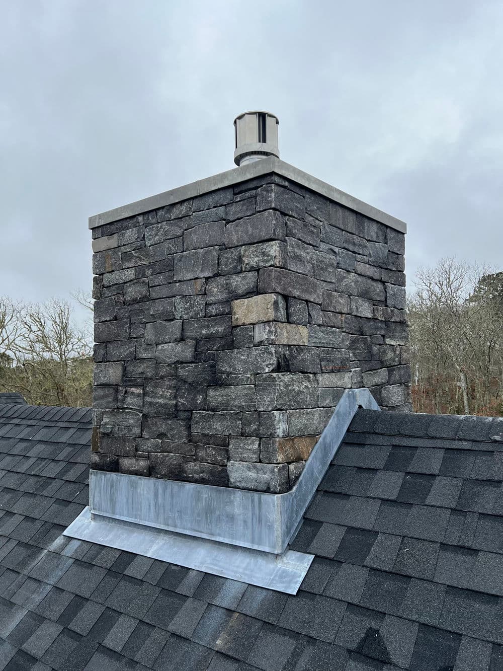 Stone chimney atop a black shingle roof with metal flashing, under a cloudy sky.