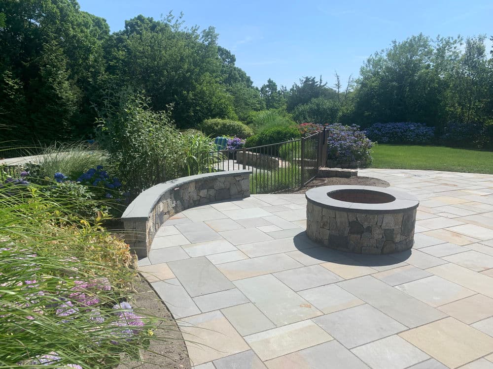 Stone patio with fire pit surrounded by lush gardens and greenery. Ideal outdoor space.